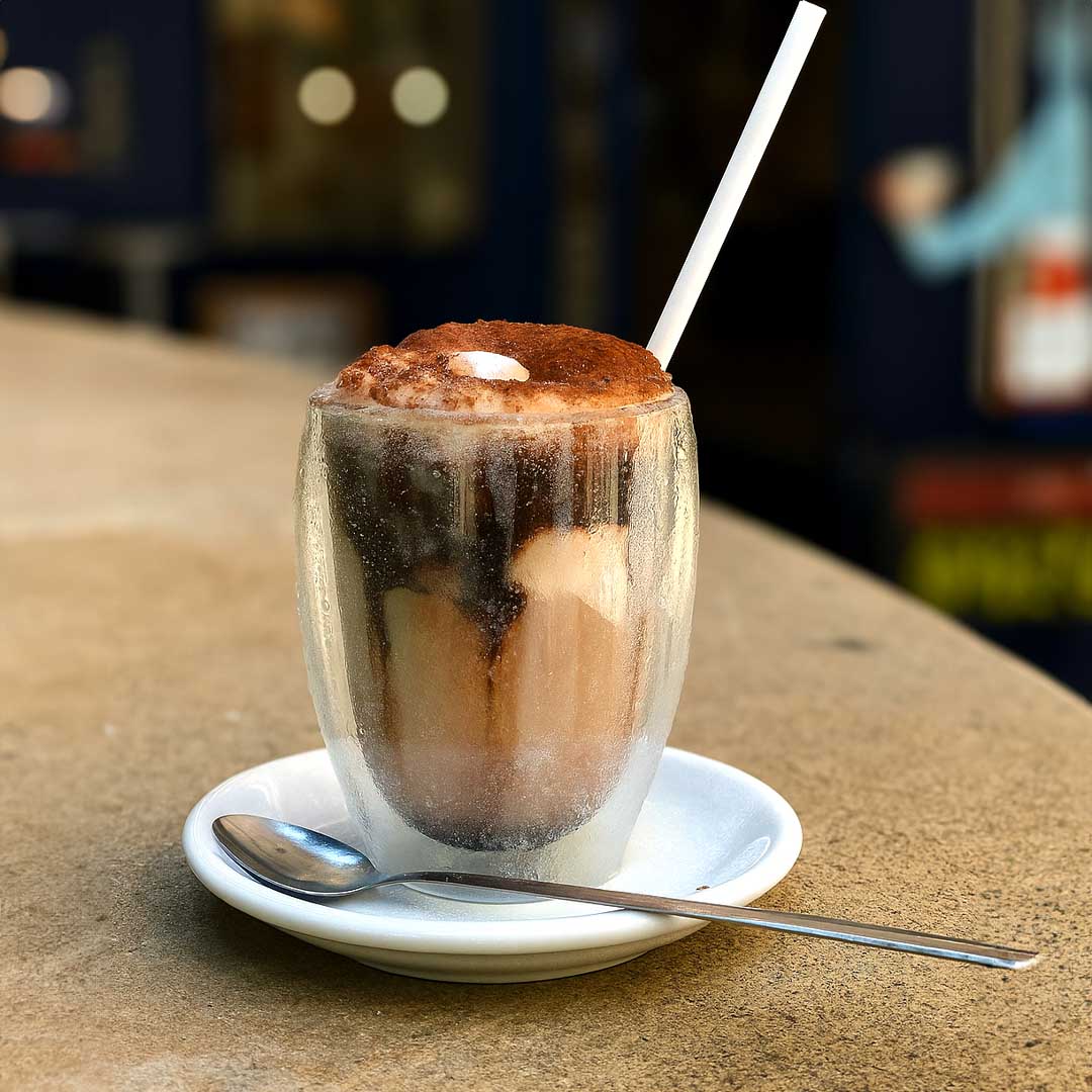 Frappuccino Cafés Maurice servi dans un verre givré, avec mousse de lait, cacao saupoudré, et paille blanche sur une terrasse d’été.